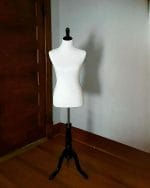A white tailor's dress form on a black stand positioned in a room with wooden flooring.