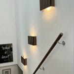 Three wooden wall lights mounted on a white wall beside a staircase with a handrail.